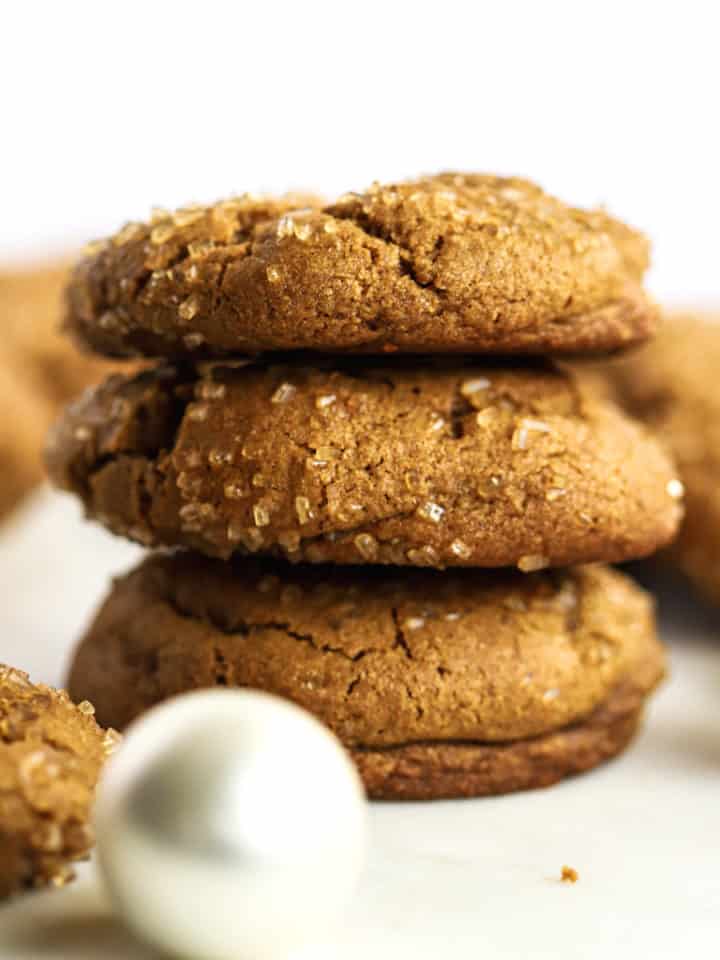 A stack of three healthier soft ginger molasses cookies, with more cookies in the background, on white marble surface with a few ornaments