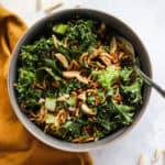 A serving of healthy Asian slaw with ramen in dark gray bowl with fork, next to mustard-colored linen on light blue and white surface