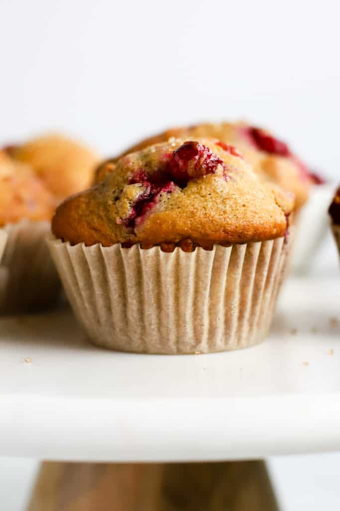 Healthy cranberry orange muffin with parchment liner on white marble cake stand, with other muffins in background