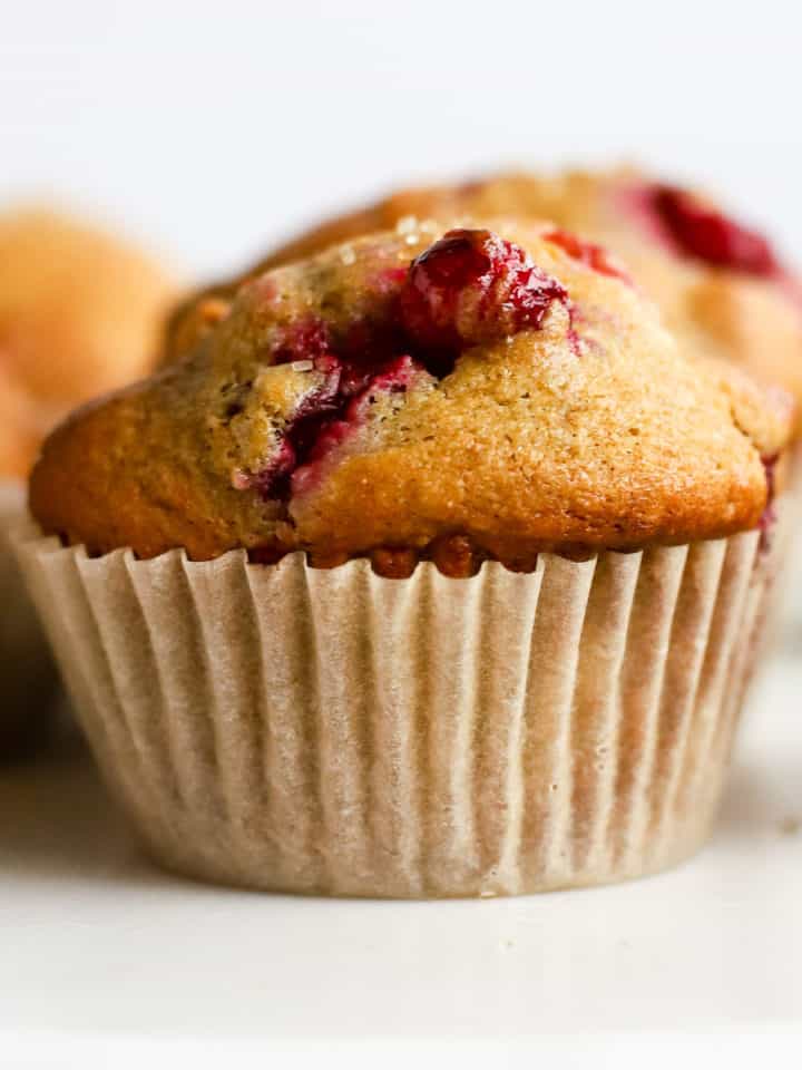 Healthy cranberry orange muffin with parchment liner on white marble cake stand, with other muffins in background