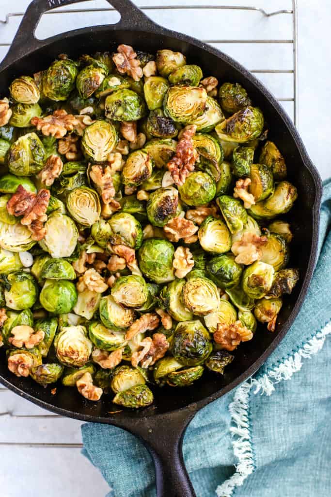 Maple walnut roasted Brussels sprouts in black cast iron skillet, on cooling rack with teal linen and light blue and white background