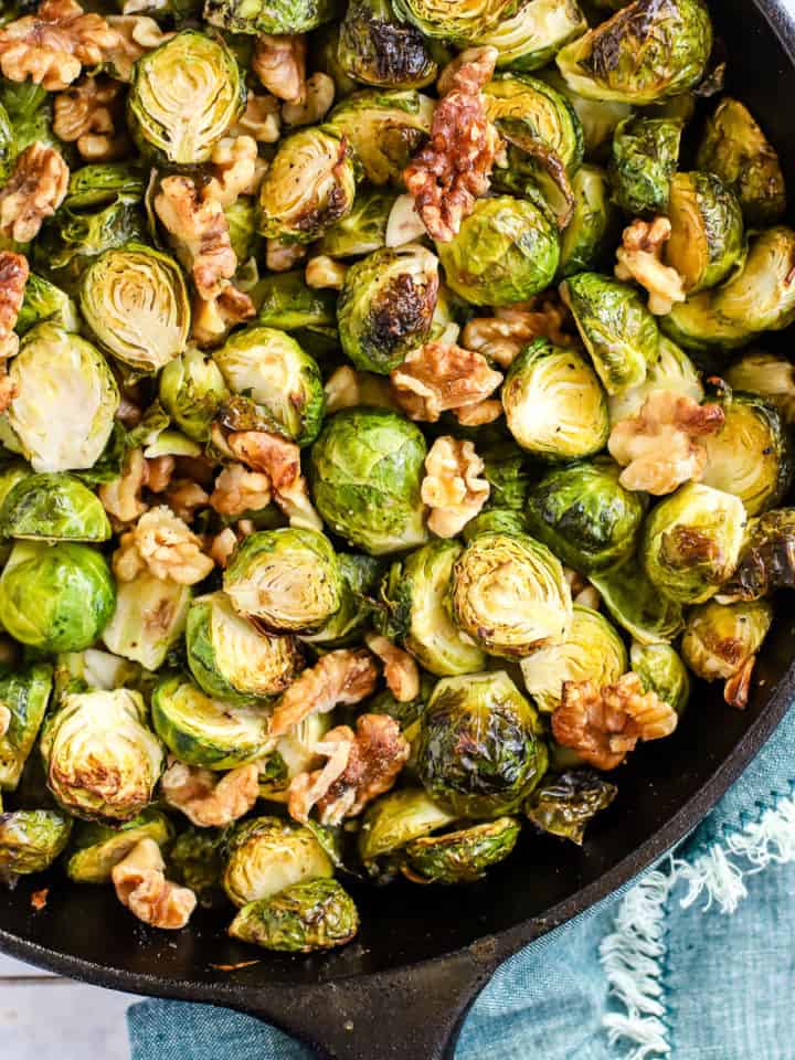 Maple walnut roasted Brussels sprouts in black cast iron skillet, on cooling rack with teal linen and light blue and white background