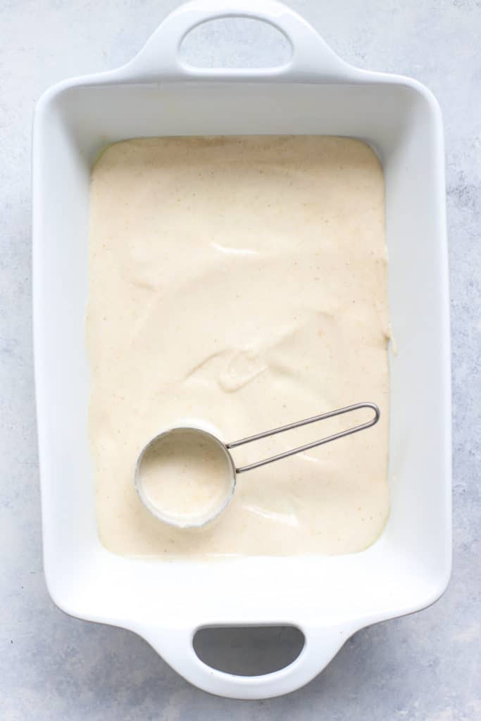 White rectangular baking sheet with white enchilada cheese sauce spread in an even layer across the bottom, on light blue and white surface