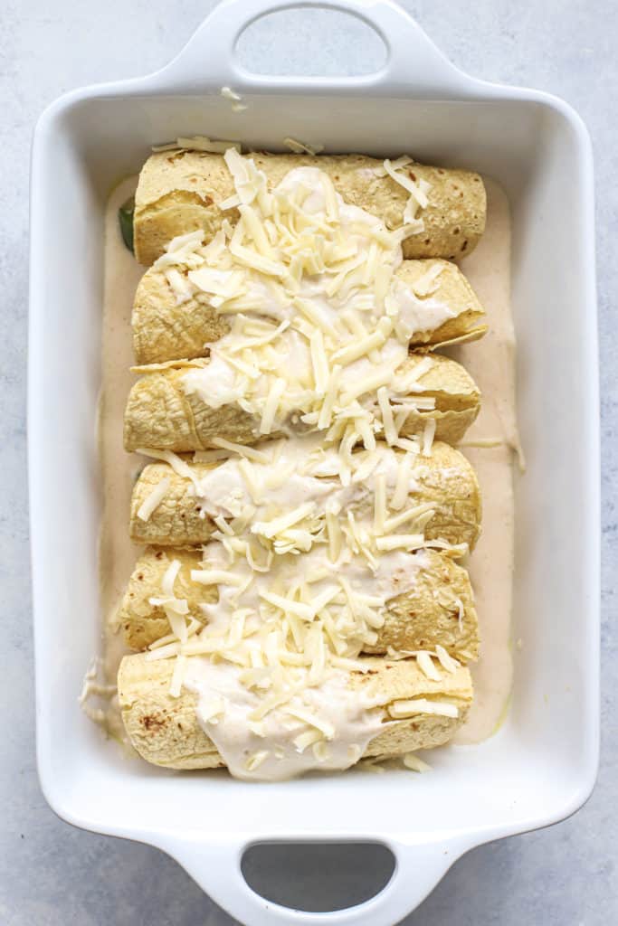 White enchiladas in rectangular white baking dish with sauce and shredded cheese on top, before going into oven