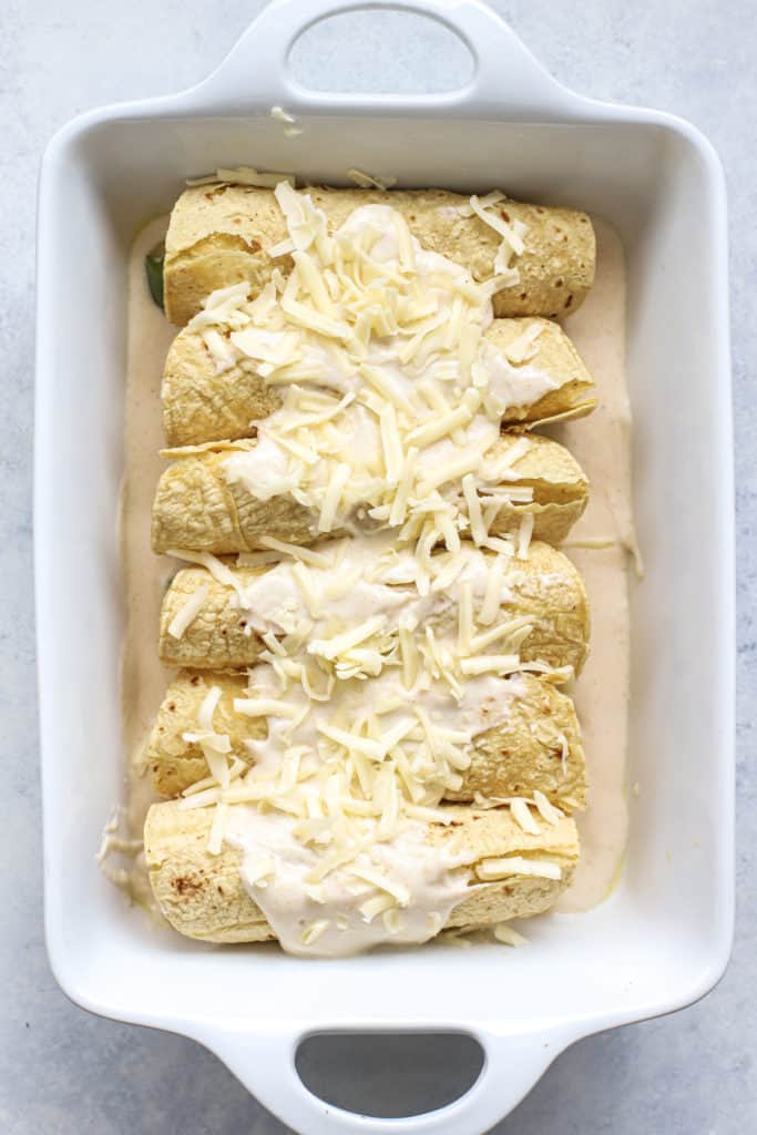 White enchiladas in baking dish with sauce and shredded cheese on top, before going into oven.