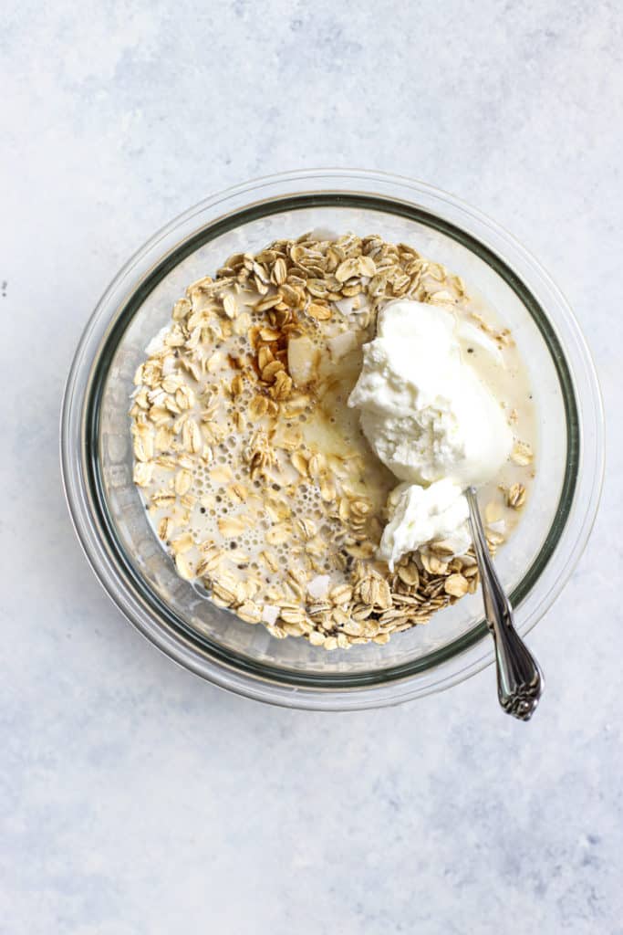 Milk and Greek yogurt added to oats, chia seeds, and coconut flakes in clear glass bowl on light blue and white surface