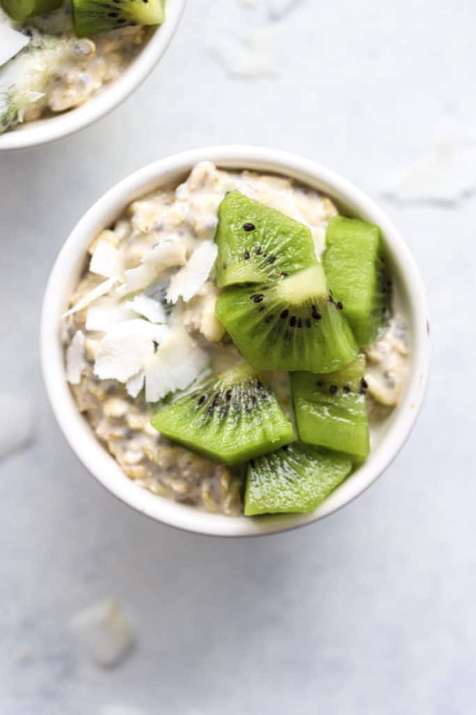 Overnight oats in small white bowl, topped with coconut flakes and diced kiwi, on white and light blue background