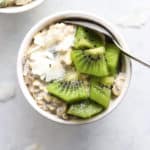 Overnight oats in small white bowl, topped with coconut flakes and diced kiwi, on white and light blue background