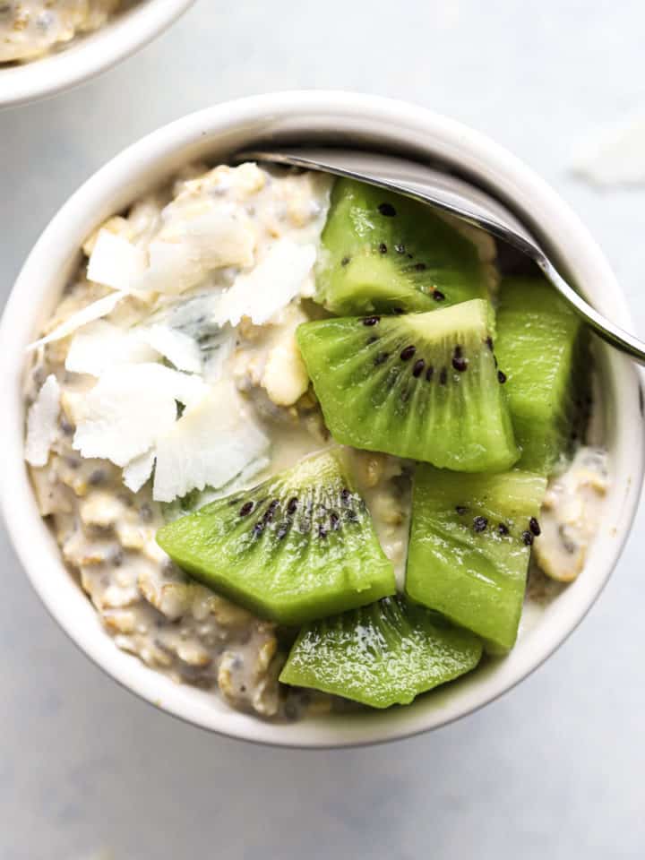 Overnight oats in small white bowl, topped with coconut flakes and diced kiwi, on white and light blue background