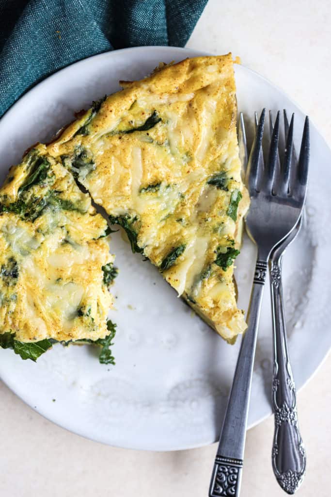 Two slices of kale, potato, and onion frittata on small white place with two forks, next to teal linen.