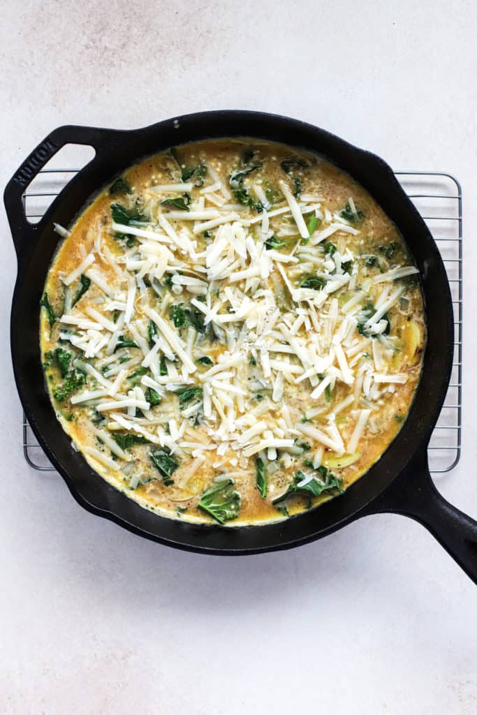 Egg mixture poured into black cast iron skillet with kale, potatoes, and onions, topped with grated cheese.