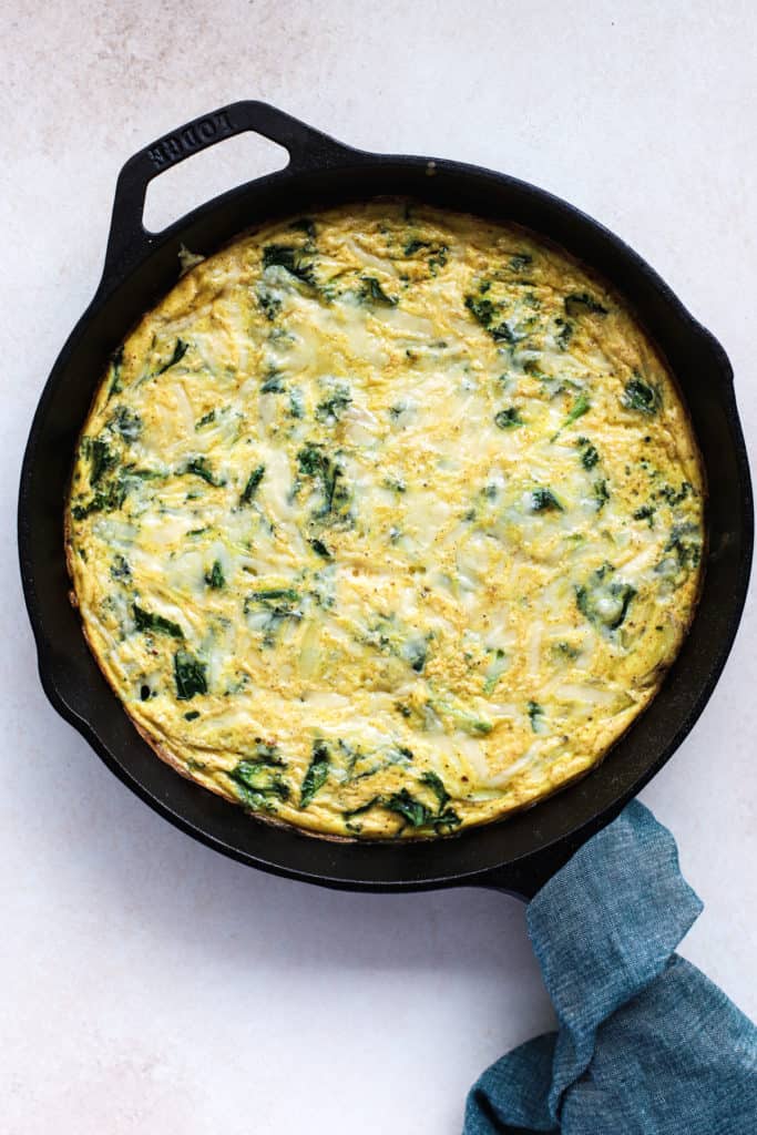 Kale, potato, and onion frittata fresh out of the oven in cast iron skillet, with teal linen wrapped around skillet handle