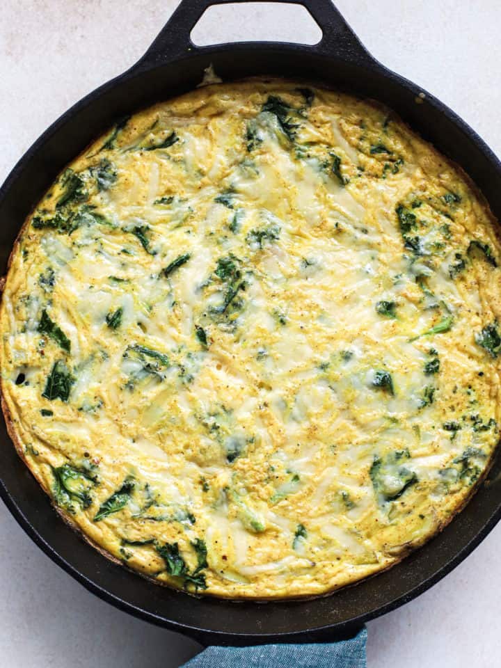 Kale, potato, and onion frittata fresh out of the oven in cast iron skillet, with teal linen wrapped around skillet handle