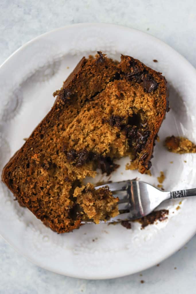Slice of spelt flour banana bread with chocolate chips on small white plate with fork, on white and light blue surface