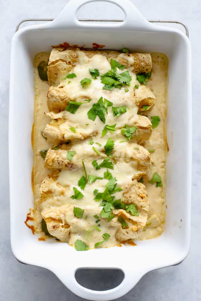 Healthy white chicken enchiladas fully baked in rectangular white baking dish. The cheese is melty and the enchiladas are topped with fresh cilantro. The baking dish sits on a wire cooling rack on blue and white surface.