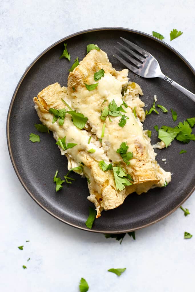 Two healthy white chicken enchiladas on dark gray plate with cilantro sprinkled on side and silver fork resting on plate.