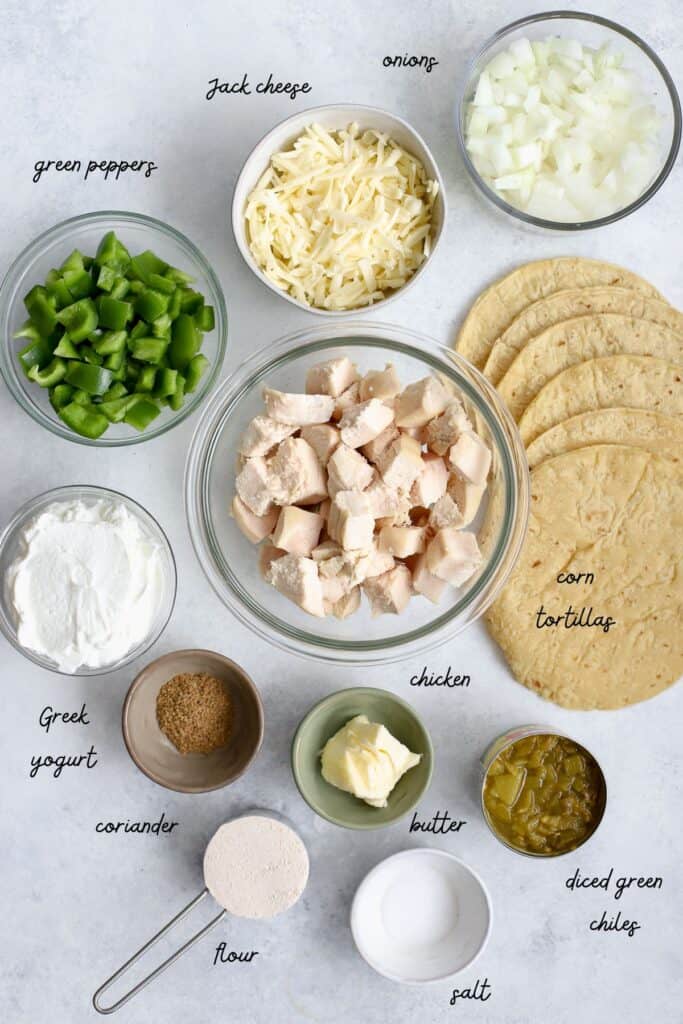 Ingredients needed for healthy white sauce enchiladas including diced green peppers, shredded Jack cheese, diced onions, whole milk Greek yogurt, chopped cooked chicken, corn tortillas, ground coriander, salt, butter, canned diced green chiles, and flour.