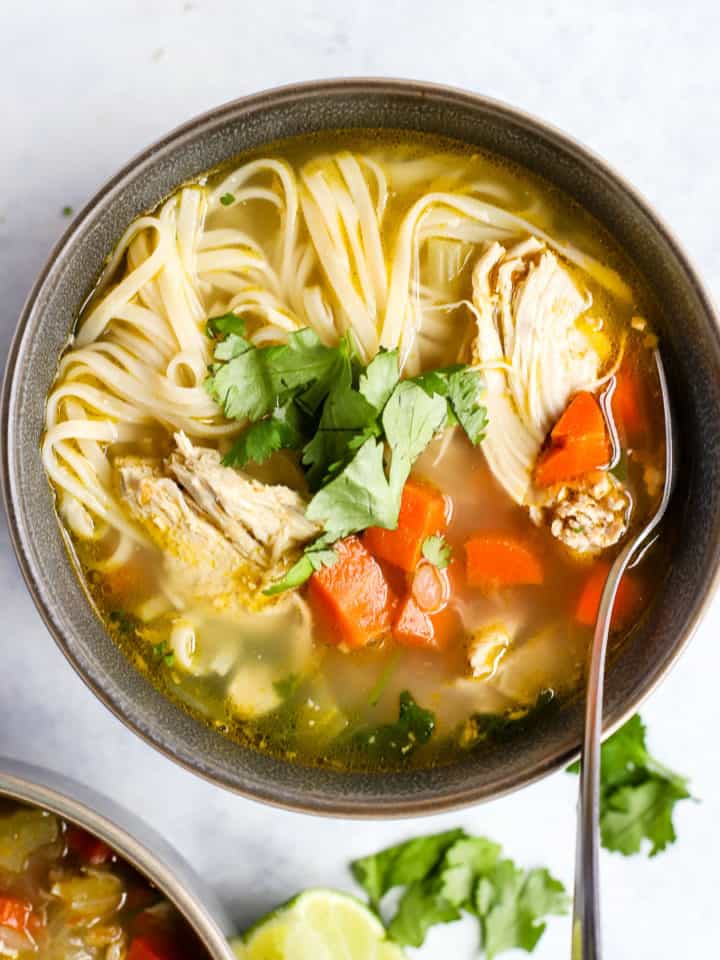 Ginger lime chicken soup with brown rice noodles, topped with cilantro, in gray bowl with spoon