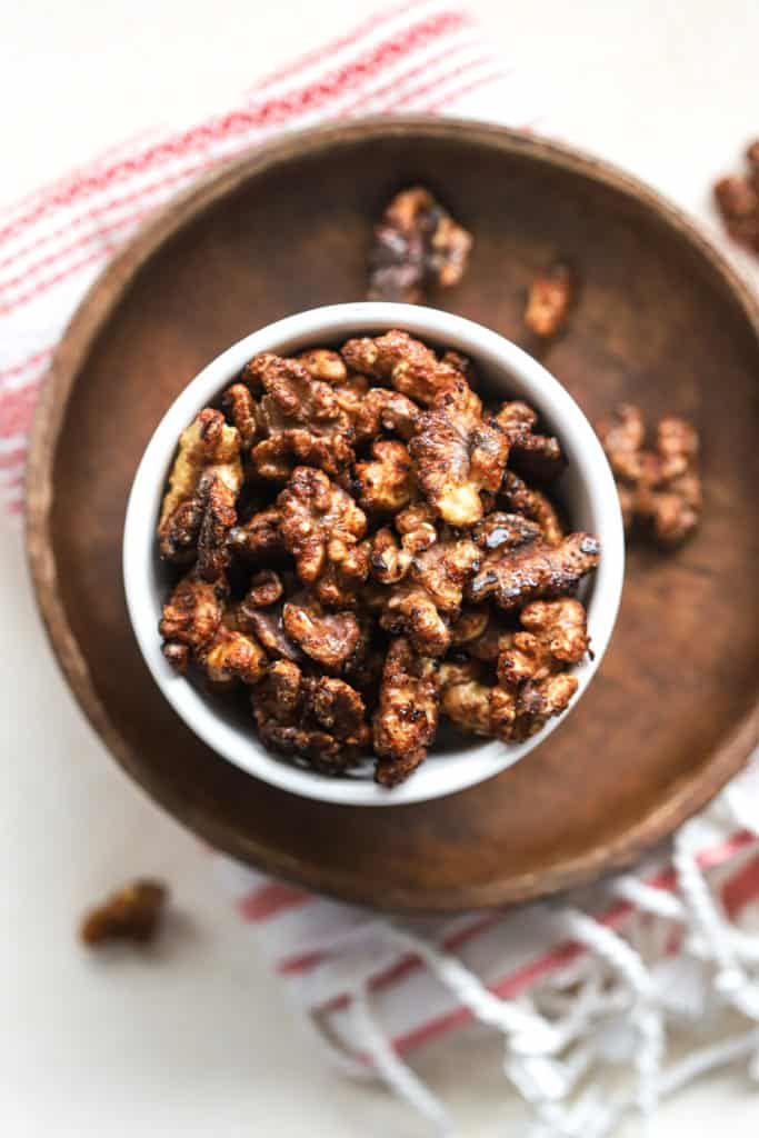 Maple cayenne toasted walnuts in small white bowl on small wooden plate with other walnuts sprinkled around, on white and pink linen