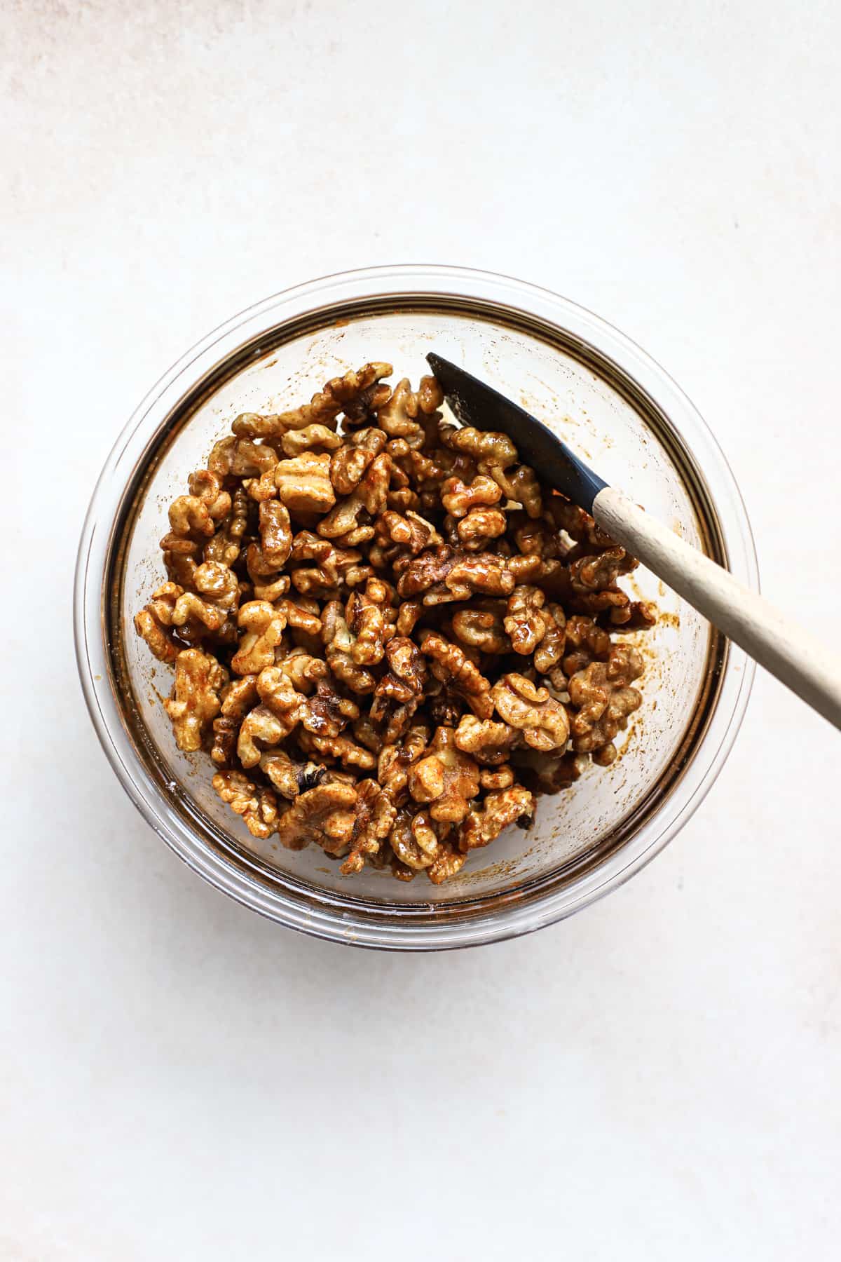 Walnuts, cayenne, salt, and maple syrup tossed together in clear glass bowl with spatula, on beige and white surface