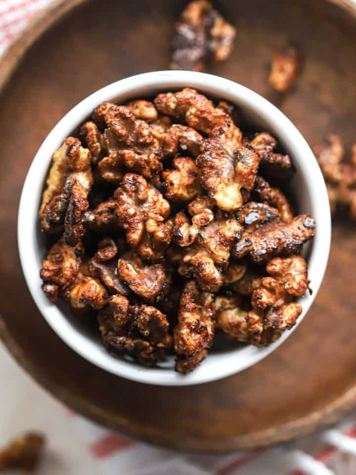 Maple cayenne toasted walnuts in small white bowl on small wooden plate with other walnuts sprinkled around, on white and pink linen