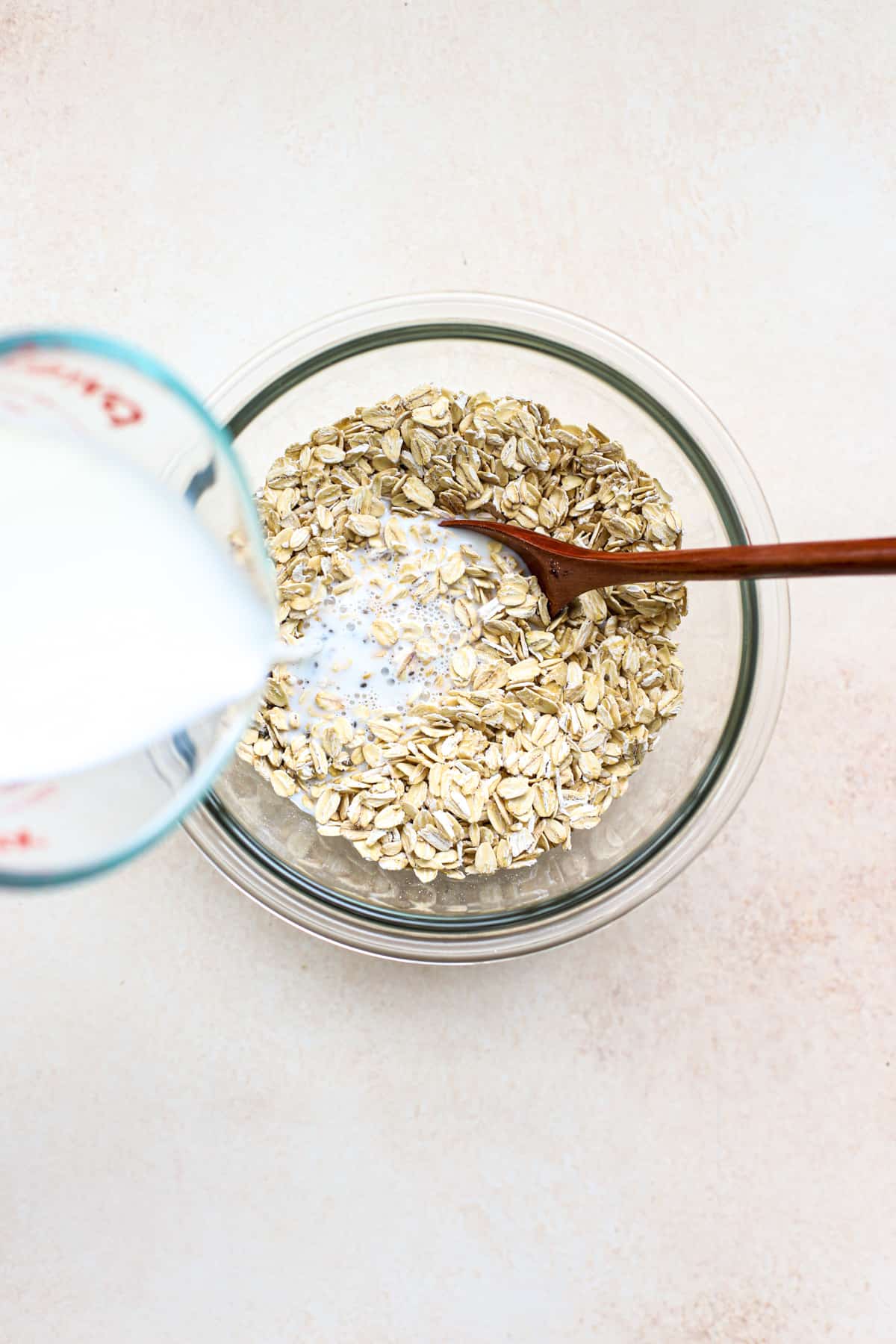 Milk pouring into oat mixture in clear glass bowl with wooden spoon