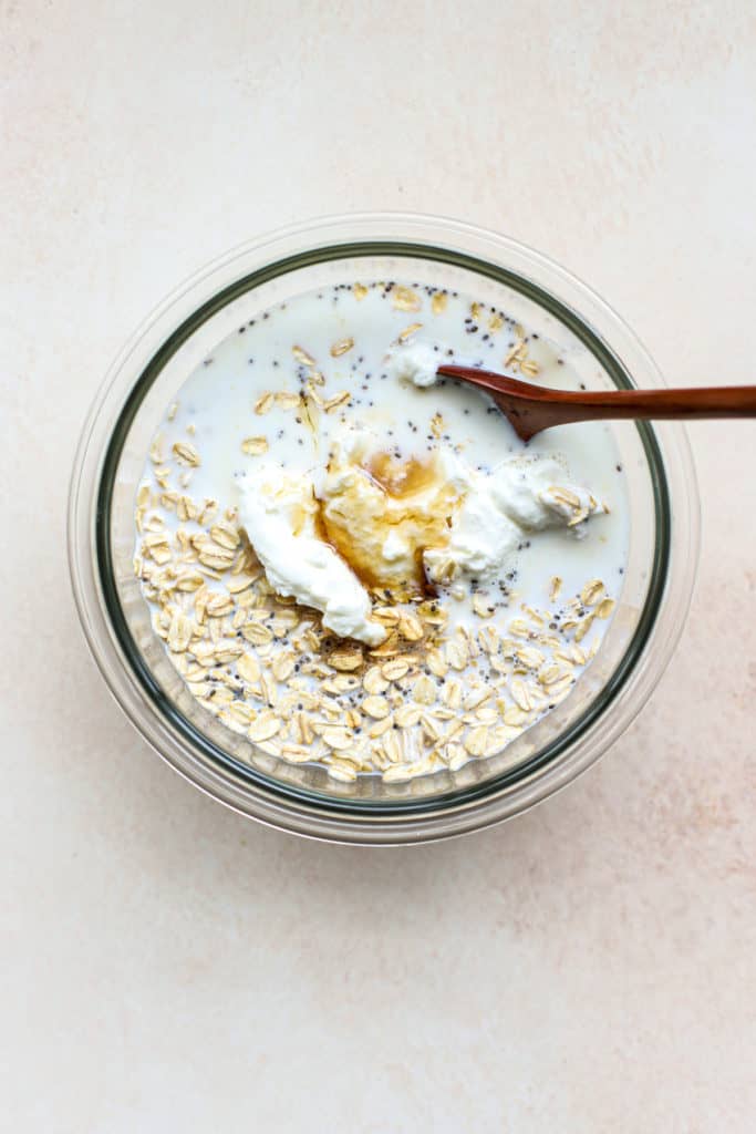 All protein overnight oats ingredients combined in a clear glass bowl before being fully stirred together, with wooden spoon.