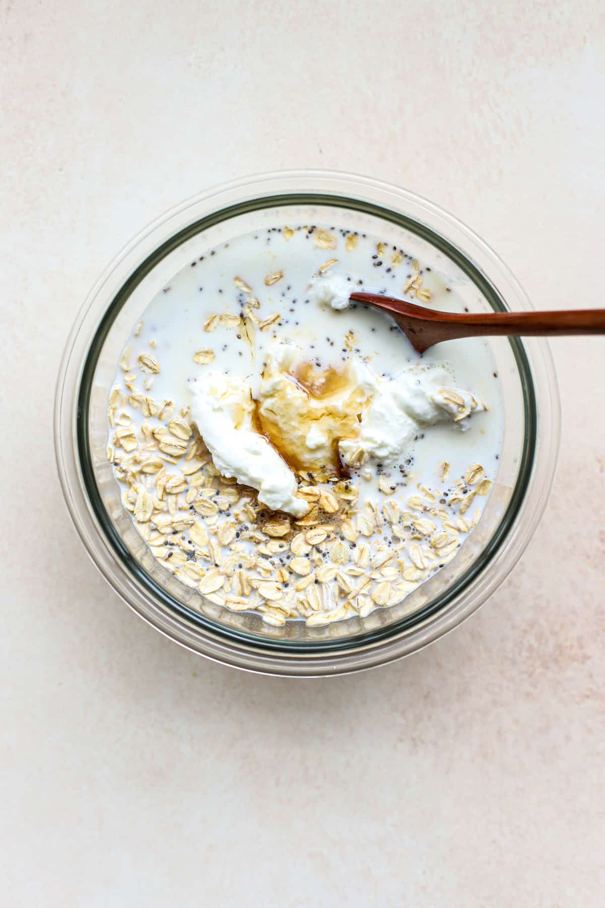 All protein overnight oats ingredients combined in a clear glass bowl before being fully stirred together, with wooden spoon