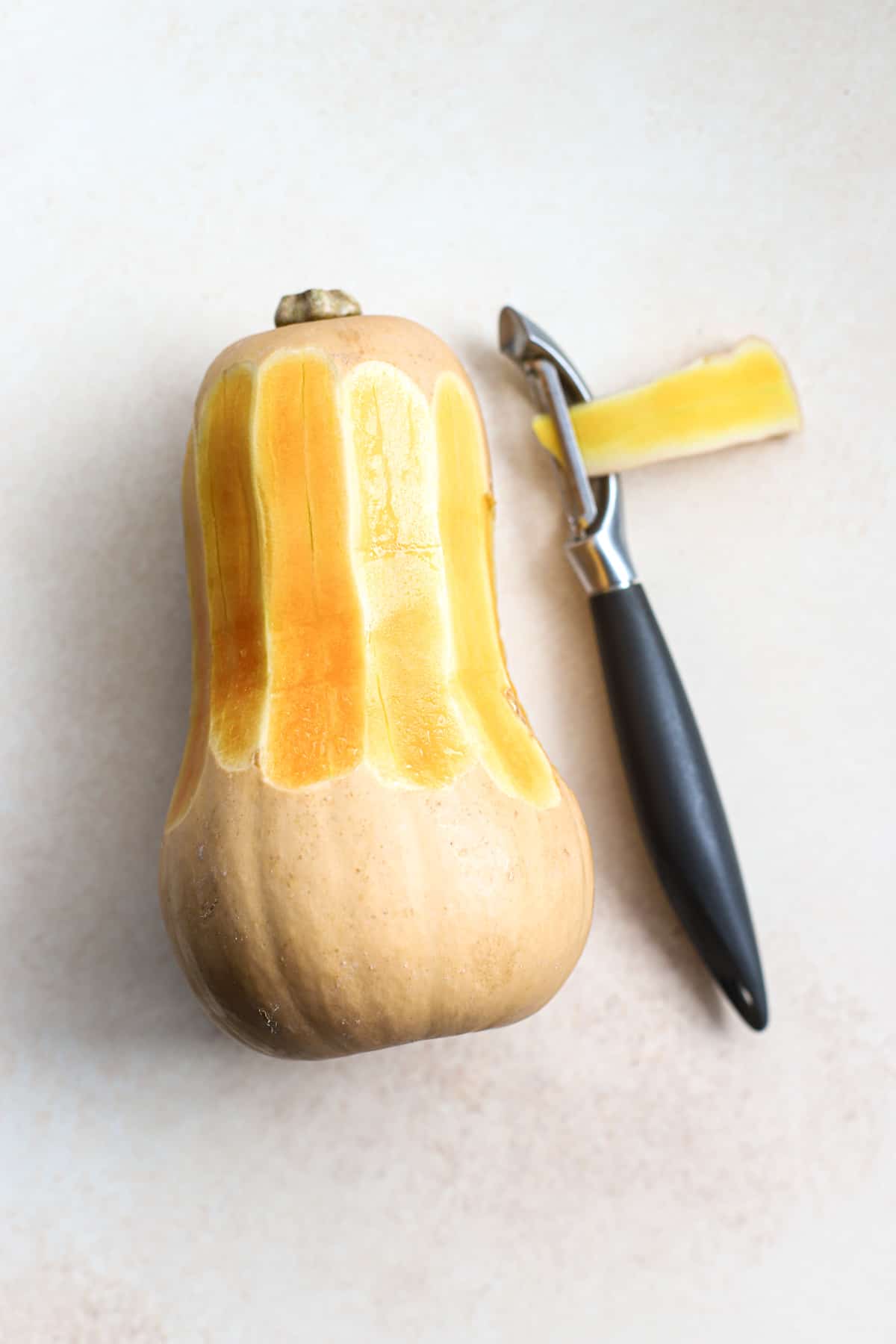 Butternut squash being peeled, next to peeler, on beige and white surface