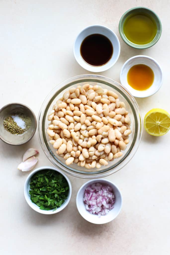 Ingredients for marinated white beans, measured out into bowls, on light beige surface