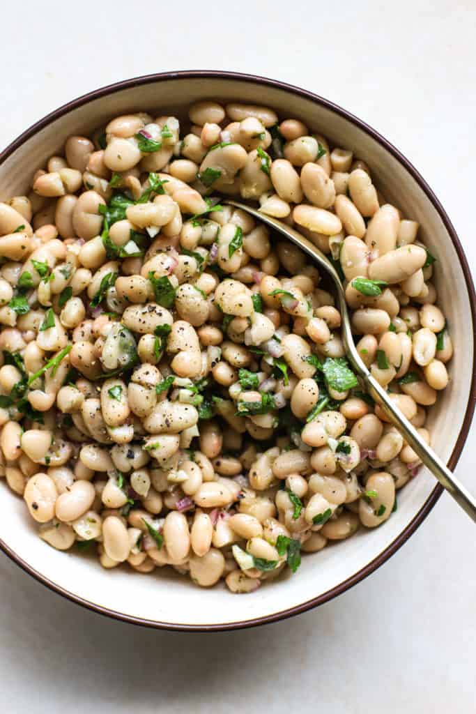 Marinated white beans in beige stoneware serving dish with golden spoon on beige and white surface