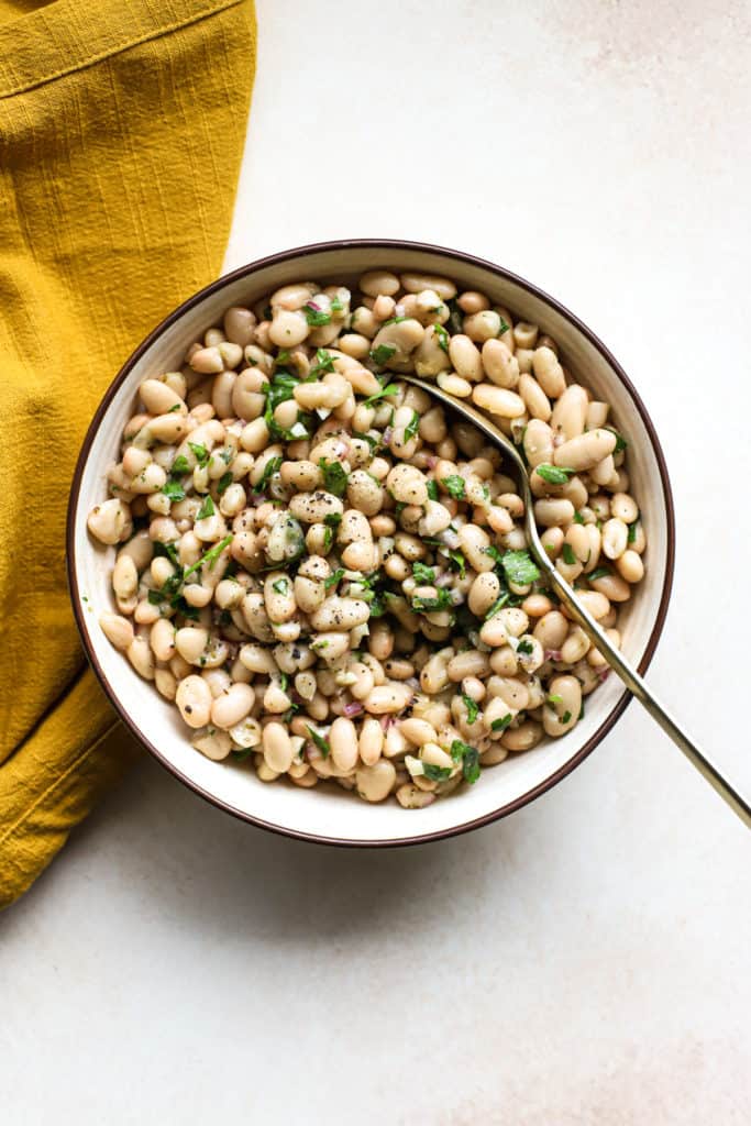 Marinated white beans in beige stoneware serving dish with golden spoon on beige and white surface, next to golden-yellow matte linen