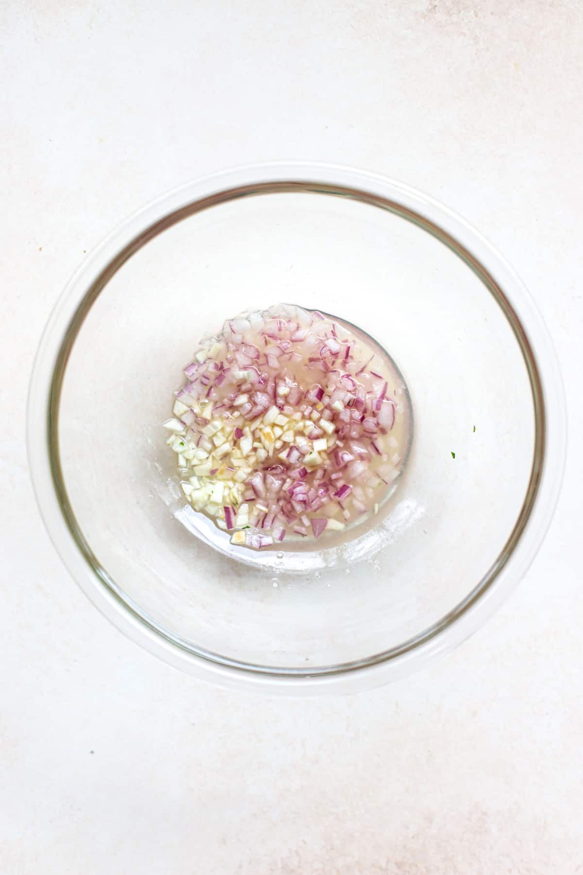 Onions and garlic macerating in lemon juice in clear glass bowl, on beige and white surface.