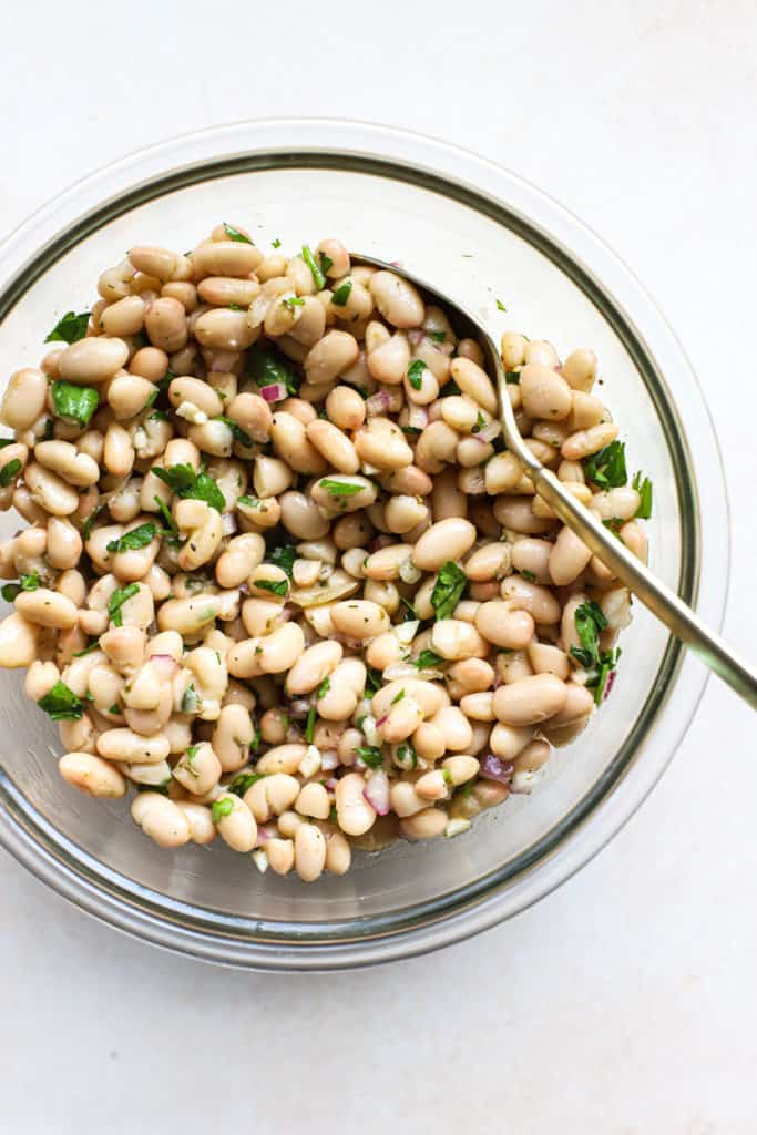 Marinated white beans in clear glass bowl with golden spoon, on beige and white surface
