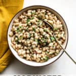 Marinated white beans in beige stoneware serving dish with golden spoon on beige and white surface, next to golden-yellow matte linen