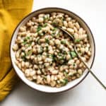 Marinated white beans in beige stoneware serving dish with golden spoon on beige and white surface, next to golden-yellow matte linen