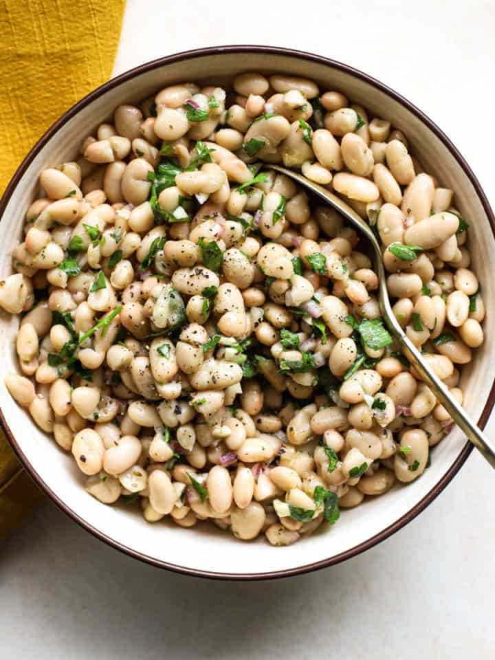 Marinated white beans in beige stoneware serving dish with golden spoon on beige and white surface, next to golden-yellow matte linen