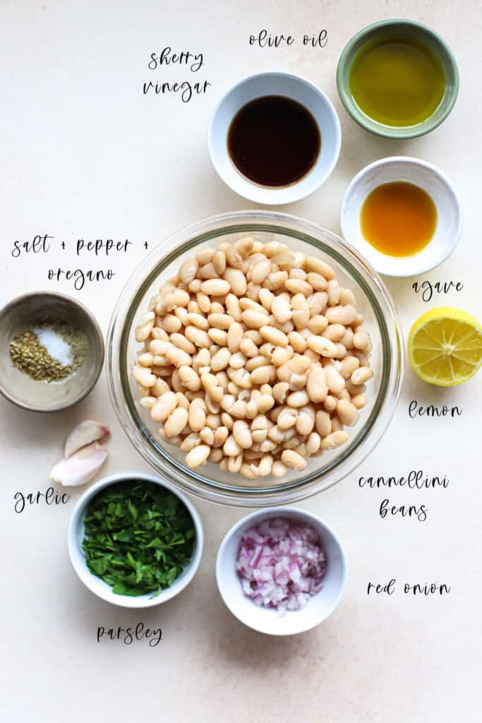 Ingredients for marinated white beans, measured out into bowls sitting on light beige surface. The ingredients include cannellini beans, olive oil, sherry vinegar, agave, lemon, salt, pepper, dried oregano, garlic cloves, fresh finely chopped parsley, and diced red onion.