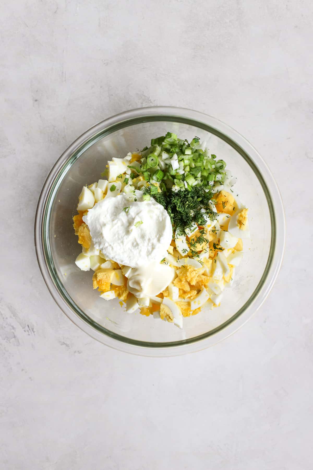 Chopped hard-boiled eggs, Greek yogurt, green onions, fresh dill, mayo, and salt in clear glass bowl on light gray surface