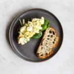 Healthy egg salad sandwich, open-faced with top slice of sourdough on the side, on dark gray plate, on light gray surface