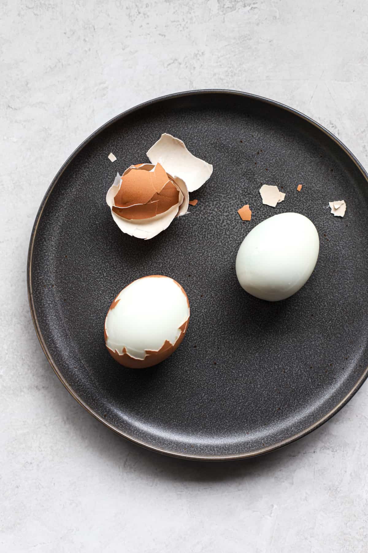 Two Ninja Foodi hard-boiled eggs being peeled easily on dark gray plate, on light gray and white surface