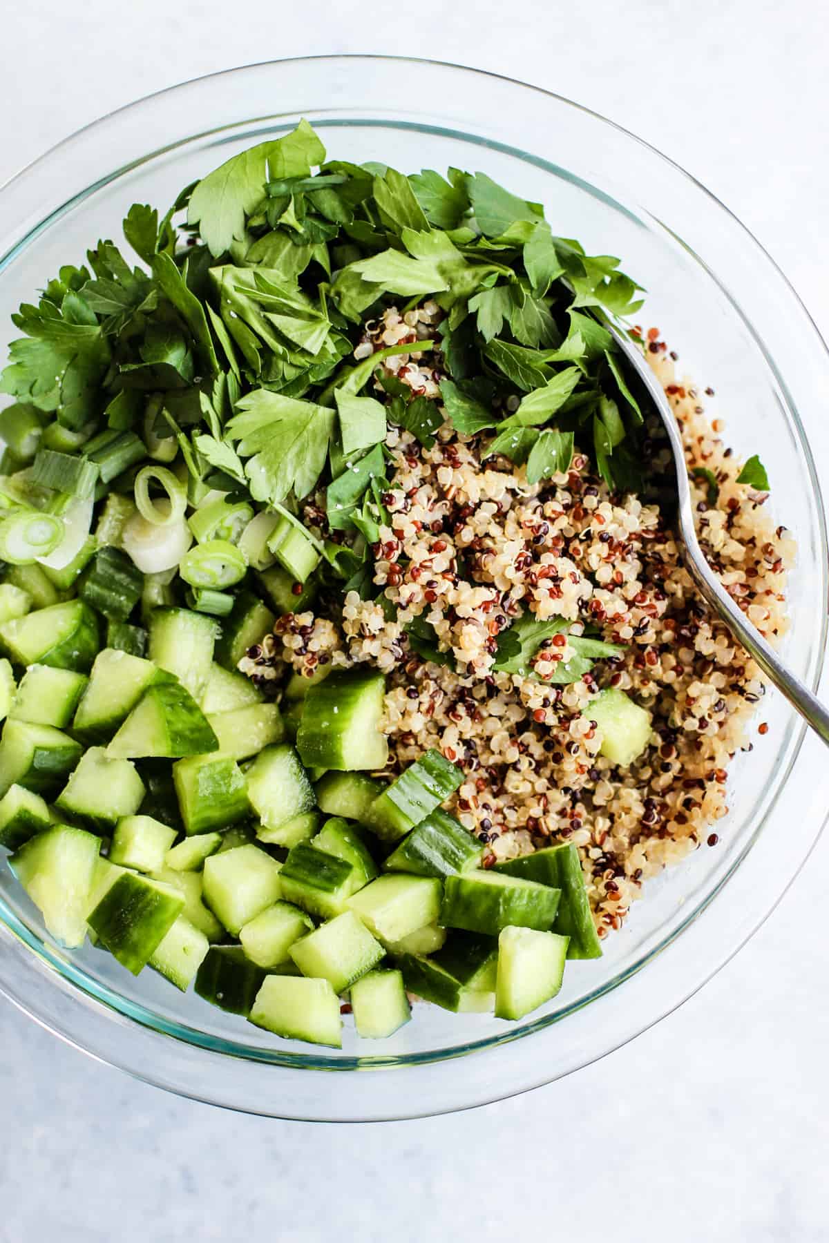 Quinoa, cucumbers, parsley, and green onions in clear glass bowl with spoon.