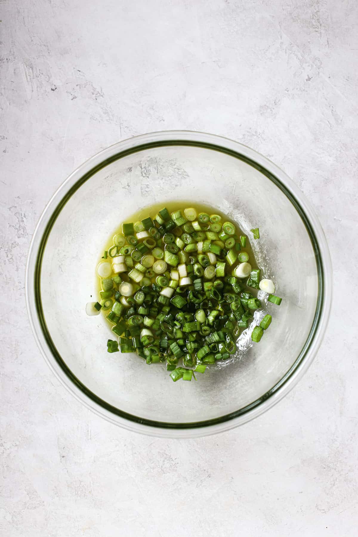 Green onions macerating in coleslaw vinaigrette in clear glass bowl on light gray surface.