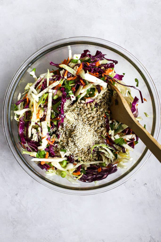 Sunflower seeds and sesame seeds poured into healthy coleslaw in clear glass bowl with wooden spoon, on light gray surface