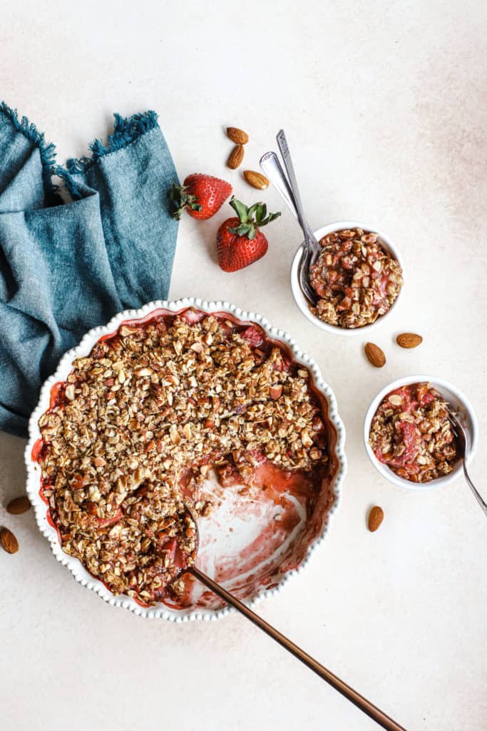 Healthy strawberry crisp in cream-colored pie dish with copper serving spoon and two small white bowls with more crisp, with almonds and fresh strawberries on the side