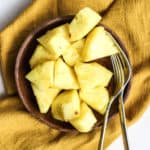 Fresh pineapple chunks on small wooden plate with two forks on gold linen, after using our favorite "how to cut a pineapple" method