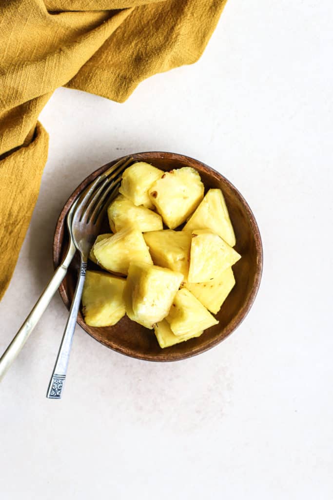 Fresh pineapple chunks on small wooden plate with two forks on beige and white surface, next to gold linen