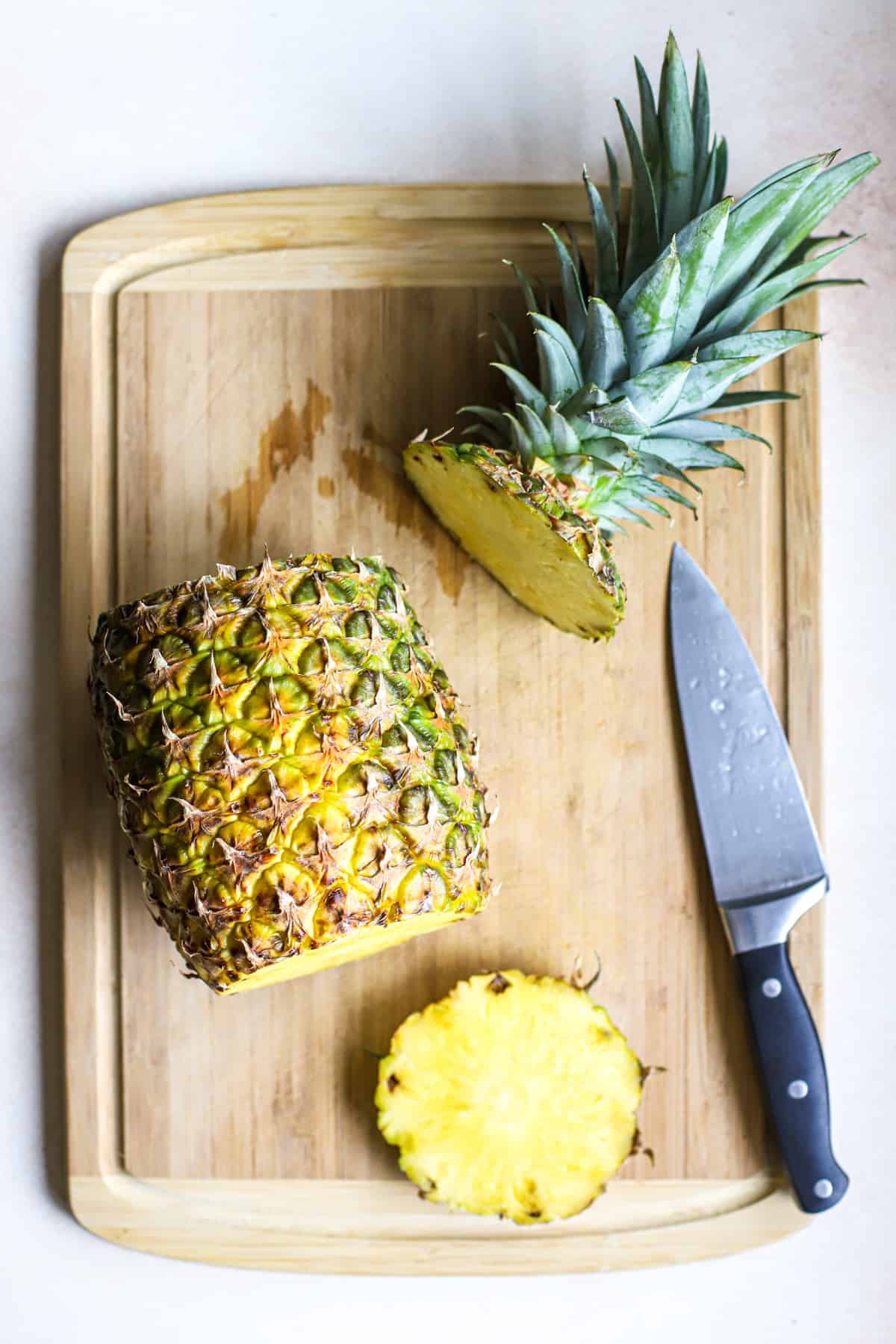 Whole fresh pineapple with top and bottom sliced off, on bamboo cutting board next to chef's knife. This is step one in "how to cut a pineapple".