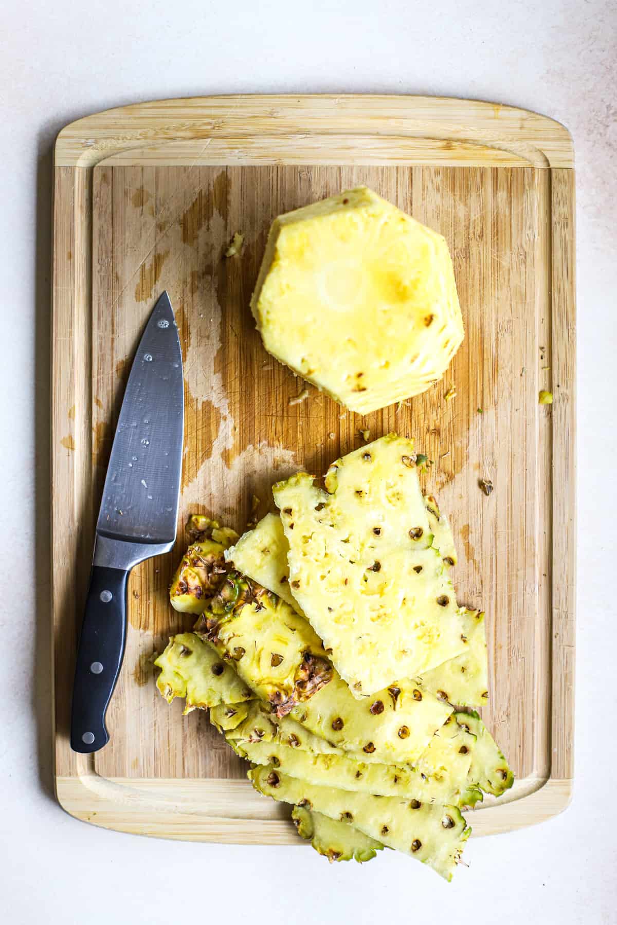 Whole fresh pineapple, peeled and stems removed, next to peels on bamboo cutting board with chef's knife