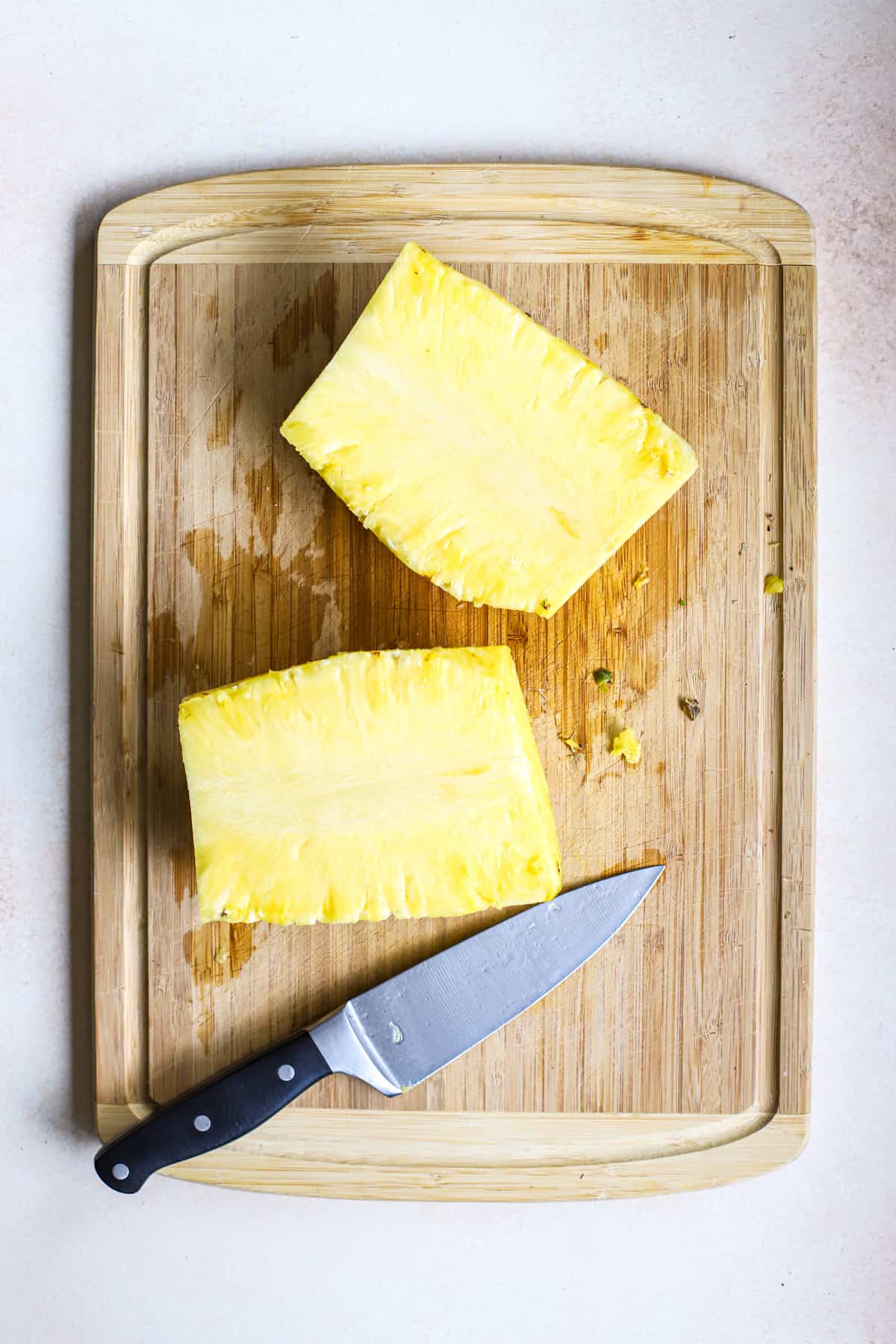 Whole pineapple cut in half, next to chef's knife on bamboo cutting board